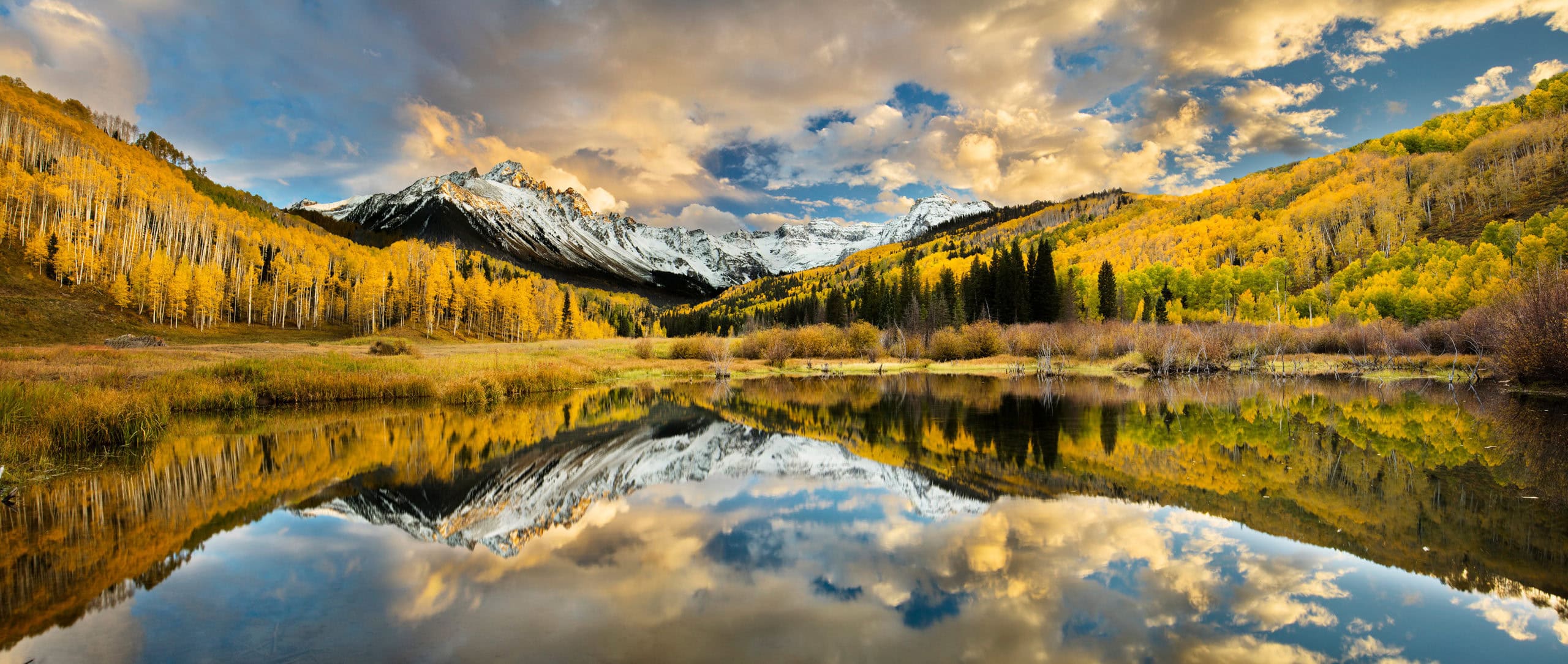 A wide panorama of peaks and cloud mirrored in still water, aspen gold running down both slopes