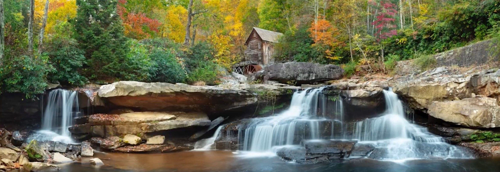 A timber grist mill standing above a wide cascade, surrounded by red and gold autumn forest