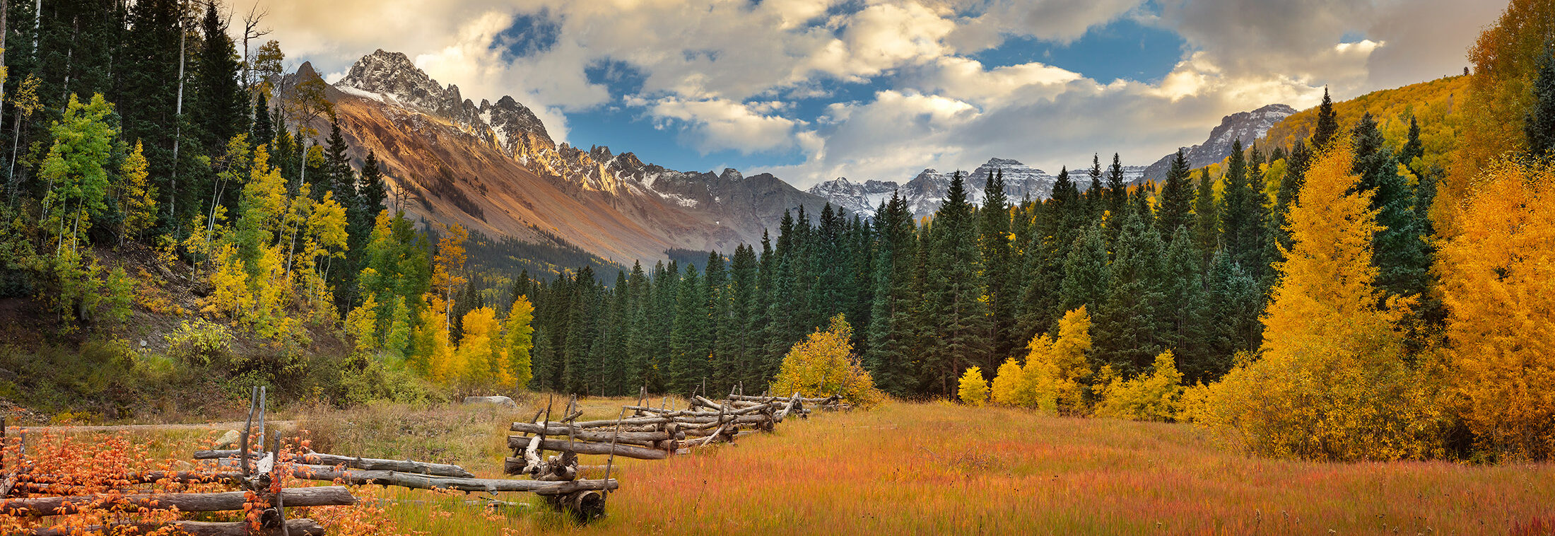 A meadow of rust-coloured grass below the San Juan peaks, split-rail fence in the foreground